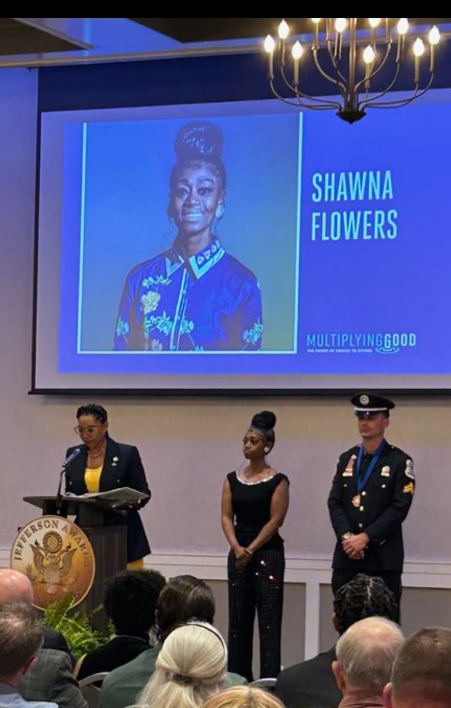Awards ceremony: speaker at podium, woman, uniformed man, and screen with Shawna Flowers' portrait.